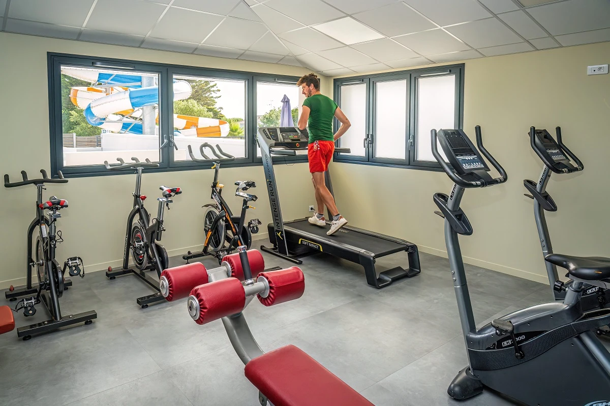 Salle de fitness du Camping Les Pirons, équipée de divers appareils de sport, dont des vélos d’appartement, un tapis de course, des vélos de spinning et un banc de musculation,