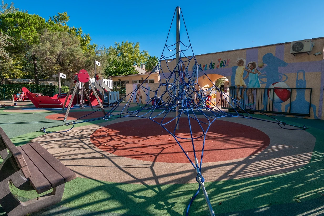 Aire de jeux colorée et sécurisée en plein air au Camping Abri de Camargue, avec une structure en toile d'araignée pour l'escalade, une aire de sol amortissant, et des éléments de jeu thématiques