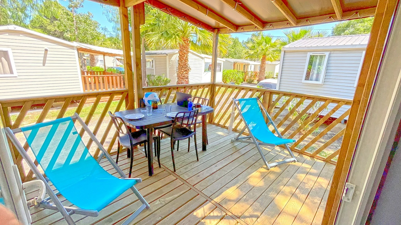  Une terrasse en bois couverte, équipée d’un salon de jardin et de transats bleus, invite à la détente et aux repas en plein air dans un cadre verdoyant, entouré de palmiers