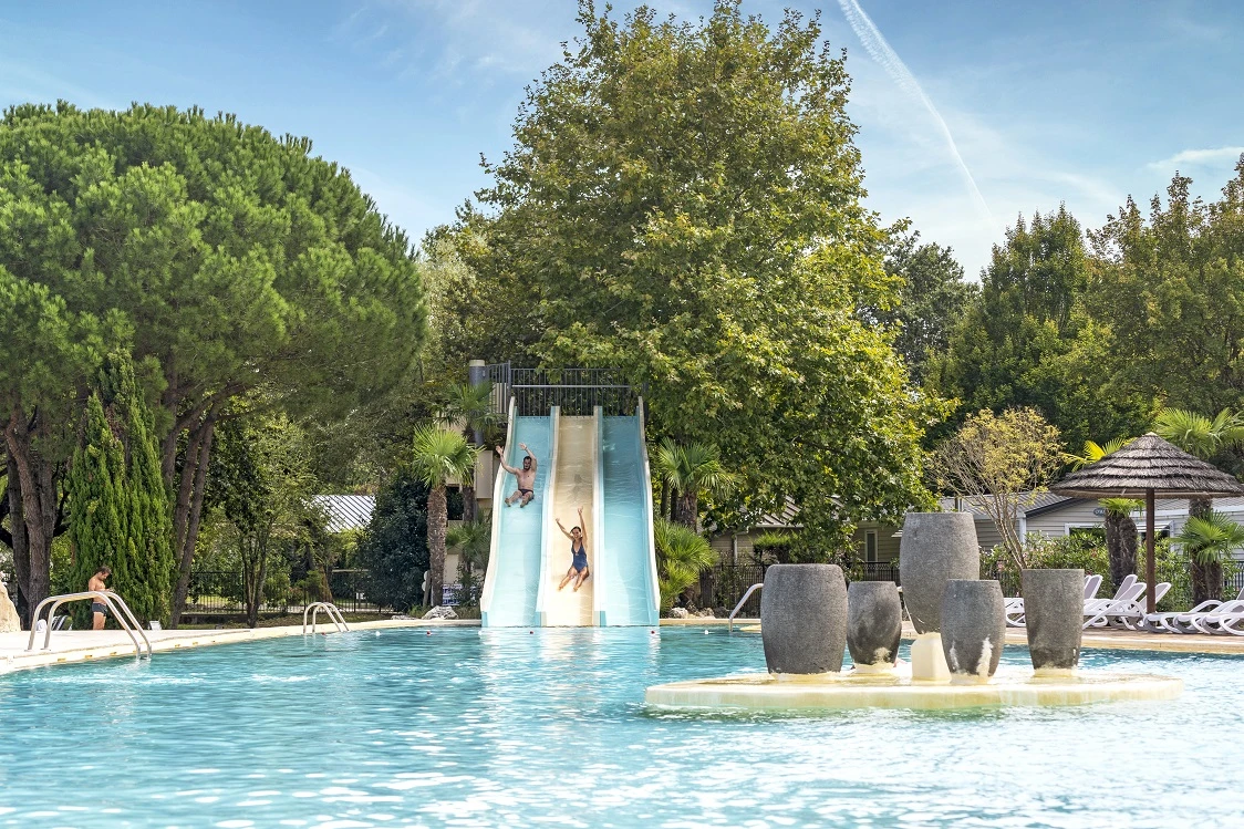 Espace aquatique lumineux et verdoyant avec une grande piscine bordée de transats et des toboggans aquatiques pour s'amuser en famille .