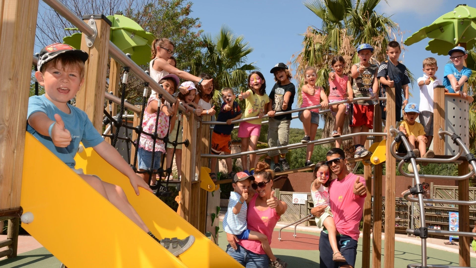 Groupe d'enfants souriants et enthousiastes jouant sur une aire de jeux en plein air dans un cadre ensoleillé, entourés d'animateurs