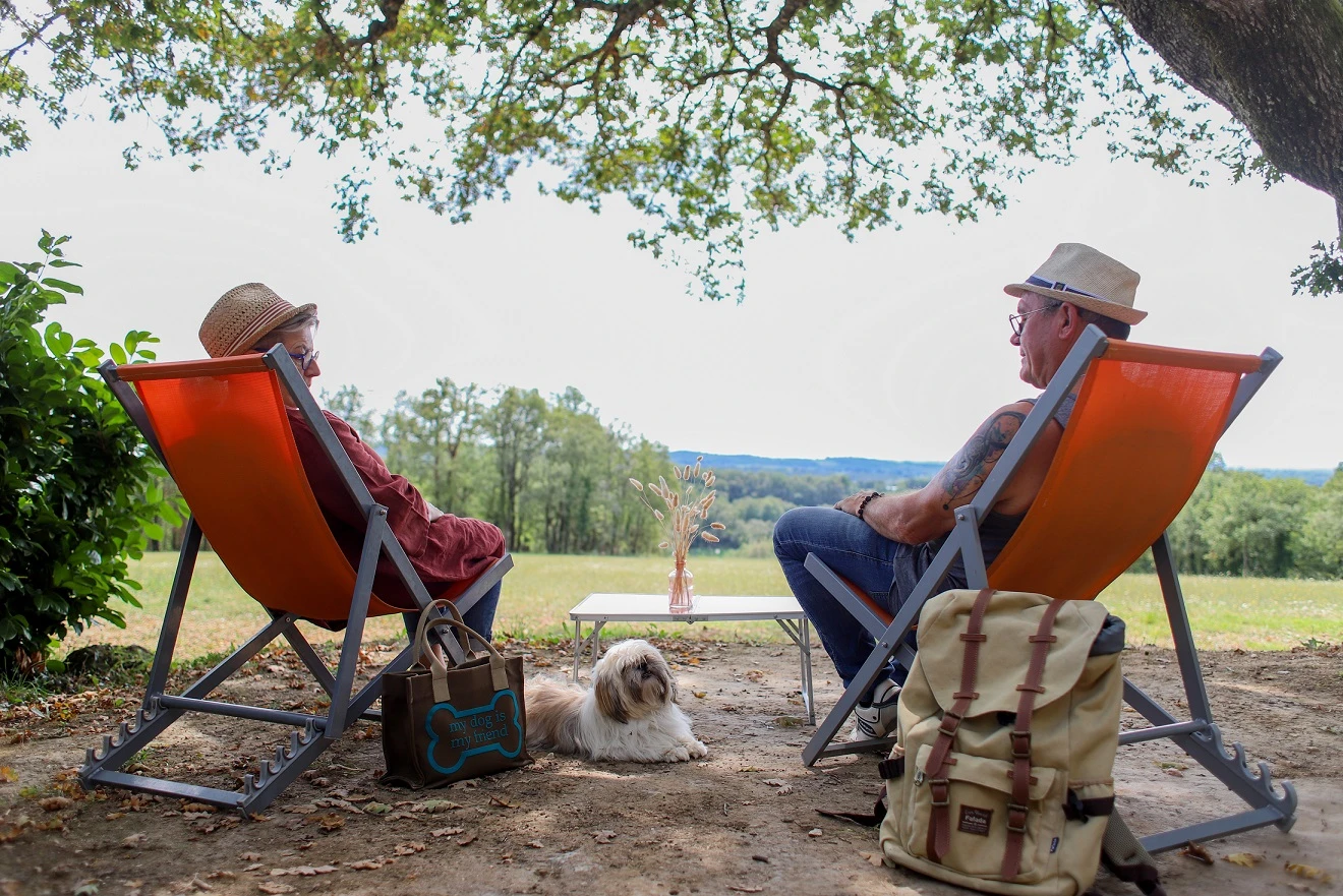  Allongé paisiblement entre ses propriétaires, ce petit chien profite d’un moment de calme en pleine nature. Installés confortablement dans des transats à l’ombre d’un grand arbre, ses maîtres admirent la vue sur les paysages verdoyants