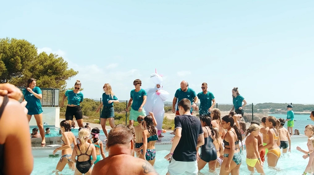 Ambiance festive dans une piscine en plein air avec des animateurs en tenue turquoise et une mascotte déguisée en licorne, entourés d'enfants au Camping L'Arquet Côte Bleue 4* 